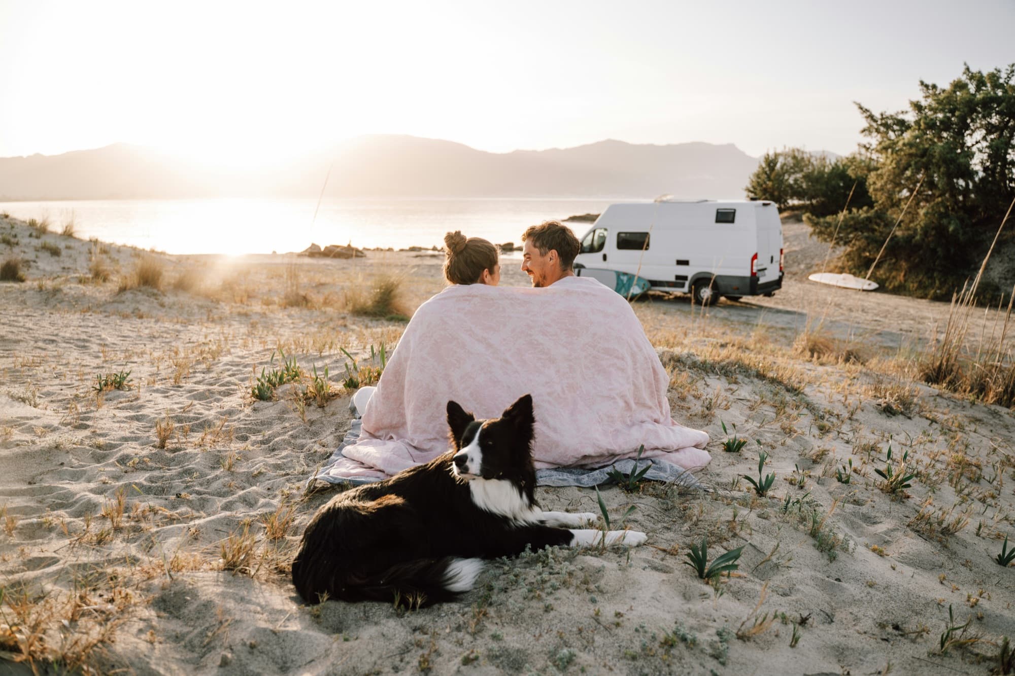 Couple enveloppé dans un plaid avec son chien face à la mer au coucher du soleil, van au loin