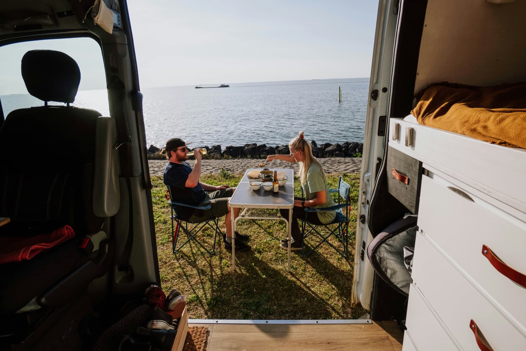 Couple partageant un repas face à la mer, portes latérales du van ouvertes