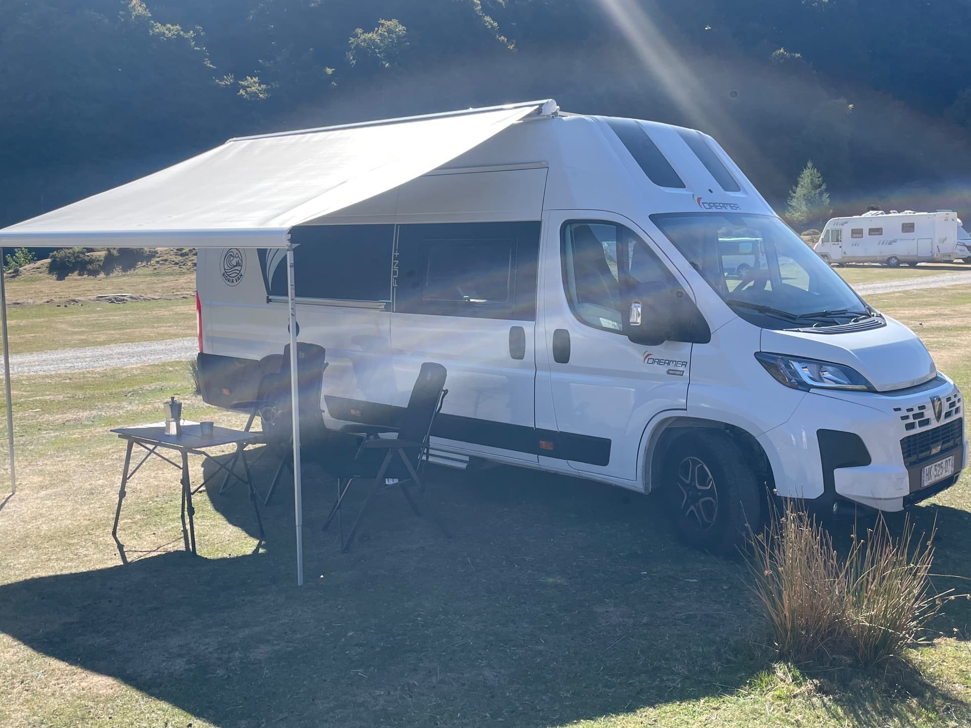 Fourgon aménagé blanc, store déployé au-dessus d'une table et de chaises sur un terrain herbeux, un camping-car en arrière-plan
